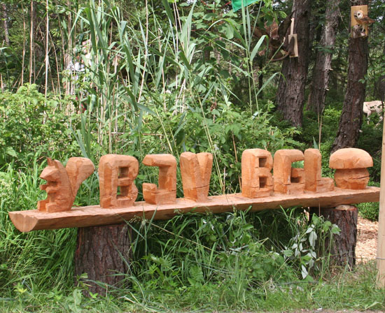 Aus Holz geschnitztes Logo des BJV. Im Hintergrund sind in Bäumen aufgehängte Tierpräparate von Schleiereule und Bussard zu sehen.