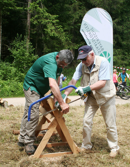 Ein Mitarbeiter der Bayerischen Staatsforsten unterstützt einen Teilnehmer beim Durchsägen eines Baumstammes.