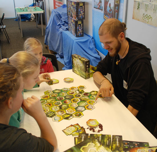 Vier junge Personen sitzen um einen Tisch, auf dem die sechseckigen Spielkarten zusammengelegt liegen. Die Personen sind nachdenklich, eine lächelt.