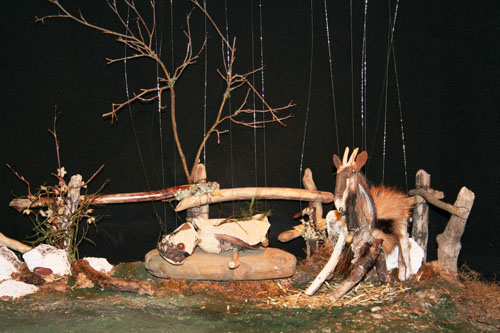Theaterkulisse: Die Bergziege Heide als Marionetten sitzt auf der Almwiese.