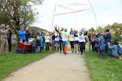 Unter einem Banner mit der Aufschrift Start läuft eine Gruppe von Leuten in weißen T-Shirts mit dem Logo des Staffellaufs. Rechts und links des Weges stehen Leute im Gras und klatschen.