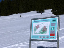 Informationstafel des DAV über die Kampagne „Skibergsteigen umweltfreundlich“, im Hintergrund zwei Skitourengeher beim Aufstieg.