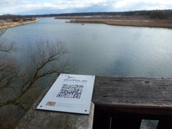 Blick von einer Brücke auf eine große Wasserfläche mit von Schilf bestandenen Halbinseln. Auf der Brüstung ist ein Schild von Ornitho.de befestigt, auf dem ein QR-Code zu erkennen ist.