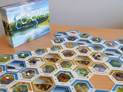  Das Bild zeigt eine Fläche aus mehreren 6-eckigen Spielelementen, im Hintergrund die aufgestellte Packung des Spieles „Ecogon – Stille Wasser“ (Foto: Melanie Schuhböck).