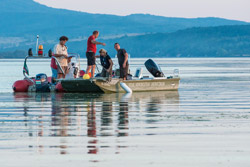 Fünf Personen stehen auf zwei nebeneinanderliegenden Booten auf einem See. Im Hintergrund bewaldete Hügel.
