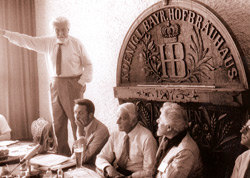 Ein Schwarz-Weiß-Foto zeigt eine Gruppe Männer, die beratend an einem Tisch sitzen, in ihrem Rücken ist das hölzerne Emblem des Münchner Hofbräuhauses zu sehen.