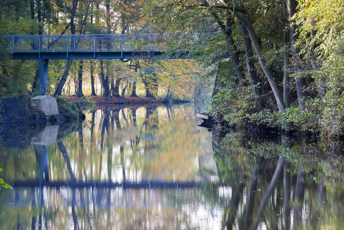 Zu sehen ist eine Brücke über einen kleinen See, der von Bäumen und Sträuchern umgeben ist. Brücke und Bäume spiegeln sich im See.