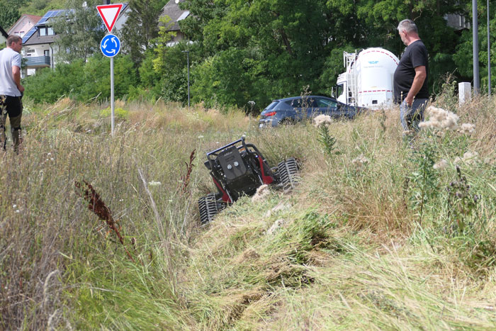 Vor einem Kreisverkehr ist zu sehen, wie sich ein Mähgerät in hohe Wiesenaufwüchse hineinarbeitet. Die Geländegängigkeit ist deutlich zu sehen, weil das Mähgerät in einem Graben in Schräglage mäht.