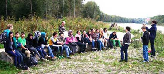 During an excursion to the Salzach students sitting on stones and listen to the speaker. In the background: The Salzach river and trees on the waterfront.