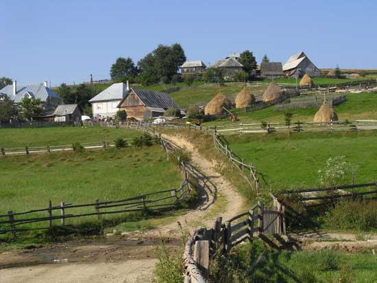 In der Mitte führt ein einfacher Weg zu höher gelegenen Häusern. Links und rechts davon grenzt die Holzzäunung die anliegenden Weiden ab.