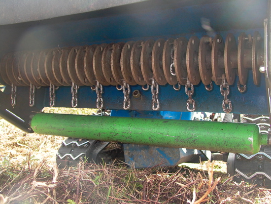 Die Unterseite eines Mulchers. Man sieht eine dicke, grüne Eisenstange und darüber starke Eisenketten, die zwischen Drehscheiben befestigt sind.