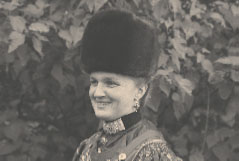 Schwarzweiß-Aufnahme; man sieht das Portrait einer lächelnden Frau, bekleidet mit einer Tracht und einer Otterhaube, einer zylinderartigen Kopfbedeckung aus Otterfell. Im Hintergrund eine Hecke.