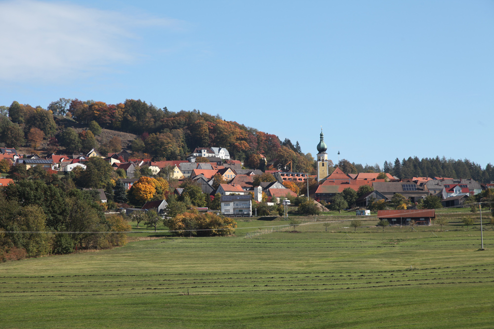 Kleines Dorf mit Kirche im Zentrum, umgeben von herbstlich gefärbtem Wald und frisch gemähten Wiesen.
