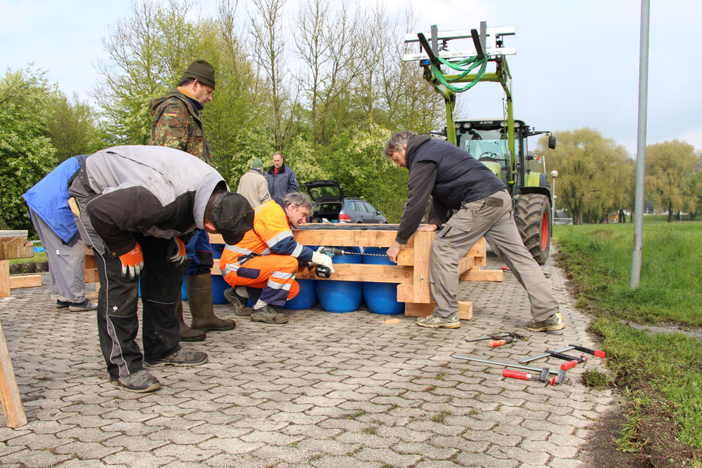 Das Bild zeigt mehrere Männer, die ein Floß montieren. Dahinter steht ein Traktor mit Gabelstapler.