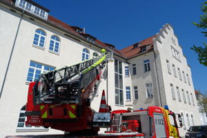 Im Vordergrund nimmt ein Feuerwehrauto mit einer Drehleiter das Bild großflächig ein. Oben auf der Leiter steht ein Feuerwehrmann. Er bringt Schwalben-Nisthilfen unter dem Dachvorsprung der sanierten Luitpoldkaserne an (Foto: Landkreis Lindau).