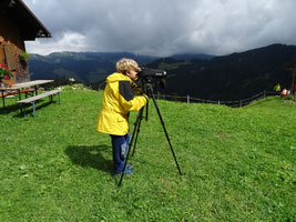 Auf einer grünen Wiese vor einer Alphütte steht ein Junge in gelbem Anorak und blickt durch ein Fernrohr ins Gebirge.