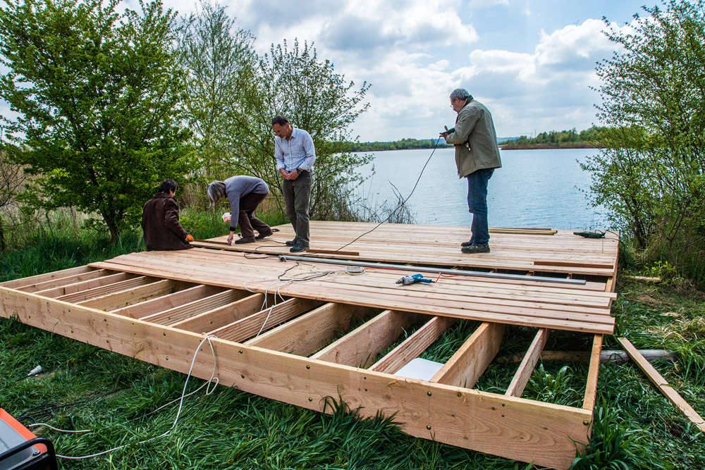 Vier Personen bauen am Ufer eines Sees ein Floß aus Holzbrettern.