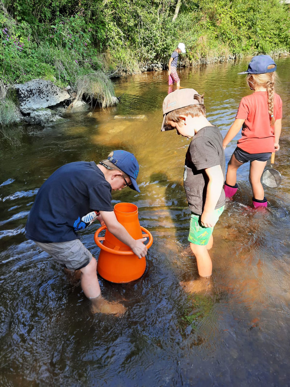 Vier Kinder stehen barfuß oder mit Gummistiefeln im Wasser, zwei der Kinder blicken durch ein Aquascope.