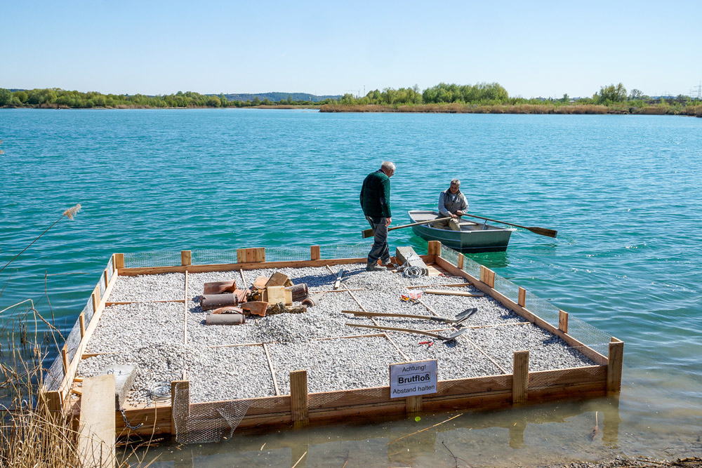 Ein Mann paddelt mit einem Boot zu einem anderen Mann, der auf einem halbfertigen Holzfloß steht.