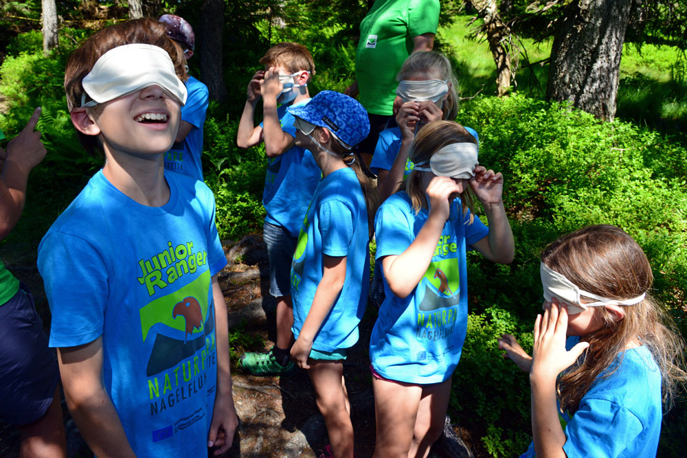 Sechs Kinder stehen in blauen T-Shirts mit dem Aufdruck „Junior Ranger“ herum und haben ihr Gesicht mit weißen Augenbinden verdeckt.