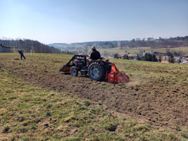 Das Foto zeigt eine hügelige Voralpenlandschaft. Im Bildvordergrund sieht man einen kleinen Traktor mit einer Fräse über eine Wiese fahren. Die Wiese ist teilweise bereits gefräst worden. Ein zweiter Mann recht im Hintergrund. Im Tal liegt ein Dorf.