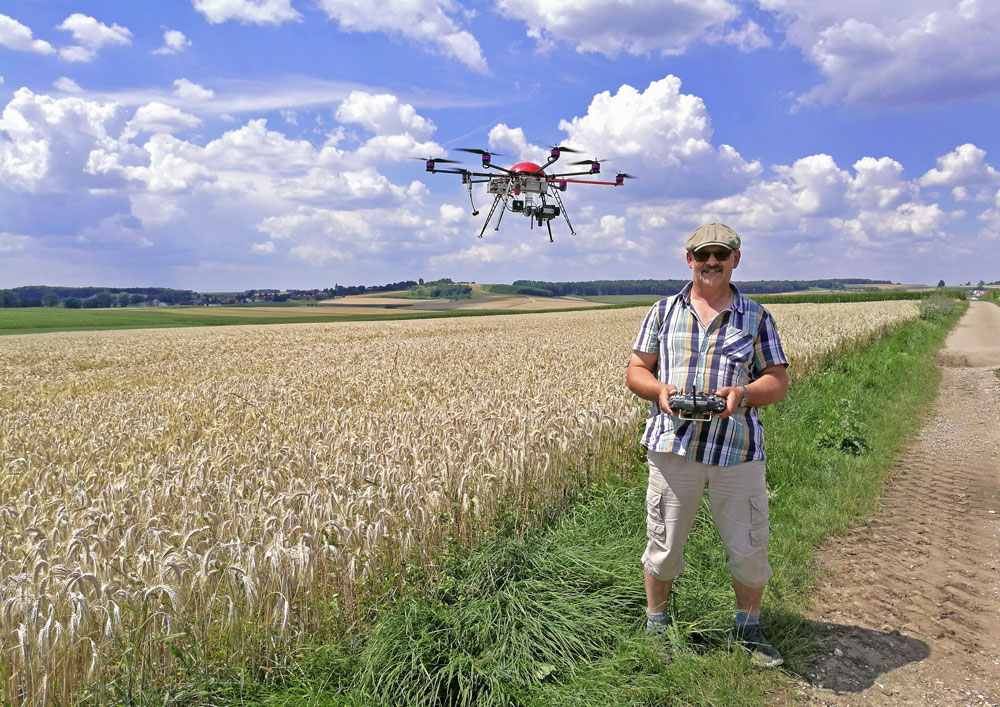 Im Zentrum des Bildes ist der Wiesenweihen-Schützer mit seiner fliegenden Drohne abgebildet. Sie befinden sich auf einem Feldweg und am Rand eines reifen Getreidefeldes. Hinter ihnen erstreckt sich die weite Tallage des Nördlinger Rieses mit ihren fruchtbaren landwirtschaftlichen Böden. 
