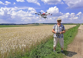 Im Zentrum des Bildes ist der Wiesenweihen-Schützer mit seiner fliegenden Drohne abgebildet. Sie befinden sich auf einem Feldweg und am Rand eines reifen Getreidefeldes. Hinter ihnen erstreckt sich die weite Tallage des Nördlinger Rieses mit ihren fruchtbaren landwirtschaftlichen Böden. 