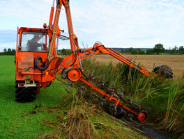 Ein orangefarbener Bagger mit speziell geformten Auslegern stutzt die Wildpflanzen an einem Feldgraben.