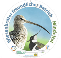 Es ist eine runde Plakette abgebildet. In farbiger Schrift trägt sie die Aufschrift „Wiesenbrüterfreundlicher Betrieb“ sowie den jeweiligen Gebietsnamen. Zentraler Bestandteil der Plakette ist eine Grafik von Kiebitz und Großem Brachvogel. Weiter sind die Logos der Beteiligten Akteure abgebildet; z. B. Landratsämter, Regierung von Schwaben oder die Landschaftspflegeverbände.