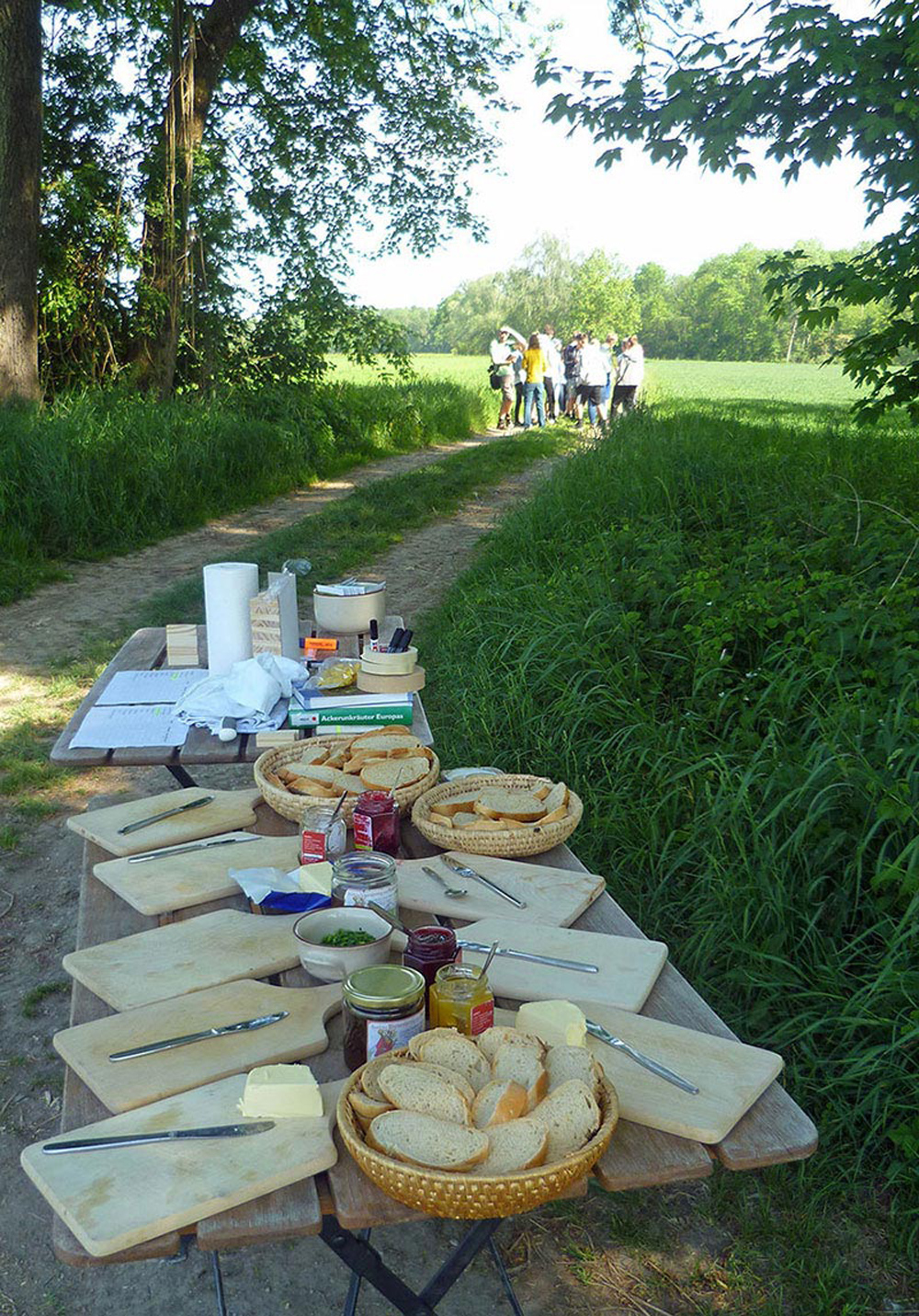 Auf einem Tisch in der freien Landschaft stehen Körbe mit aufgeschnittenem Brot und verschiedene Gläser mit Aufstrich zur Brotzeit für eine im Hintergrund wartende Gruppe von Jugendlichen bereit.