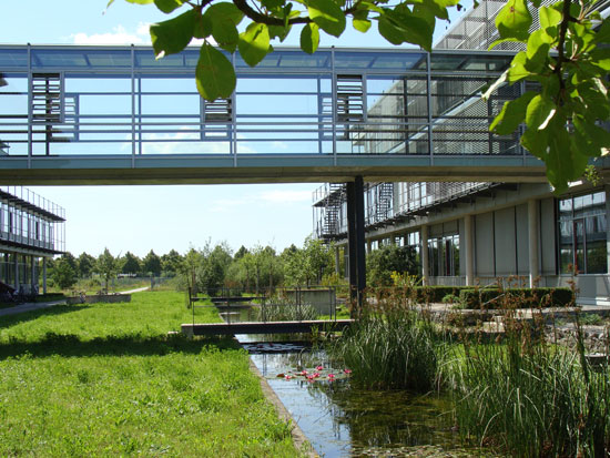 Zwischen zwei Bürogebäuden, die durch eine Fußgängerbrücke verbunden sind, liegen naturnah gestaltete Außenanlagen mit Wiesen, Bäumen und einem Kanal voller Schilf und Seerosen.