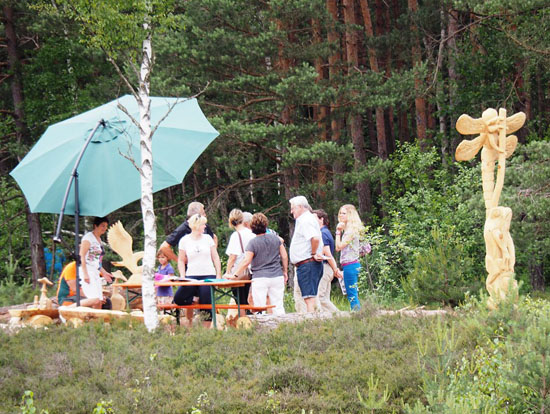 Besucher bewundern die Holzkunstwerke und beobachten den Künstler bei seiner Arbeit. Das Bild zeigt auch die Dauerleihgabe des Marterpfahls bestehend aus Schlange, Frosch und Moorlibelle.