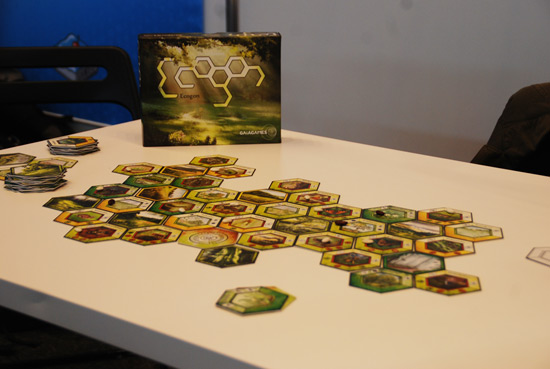 Im Vordergrund liegen die sechseckigen Spielkarten zusammengelegt auf einem Tisch, im Hintergrund stehend ist die Spielschachtel mit dem Schriftzug ECOGON zu sehen. 