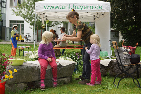 Eine Mitarbeiterin der ANL bindet mit zwei Kindern Blumenkränze.