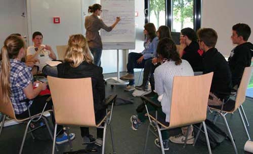 Schülerinnen und Schüler sitzen im Kreis. Eine Schülerin schreibt auf ein Flipchart.