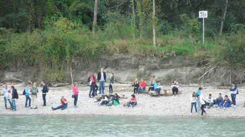 Schülerinnen und Schüler stehen und sitzen am Flussufer auf einer Sandbank.