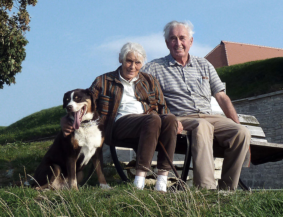 Foto eines alten Ehepaares, auf einer freistehenden Bank sitzend; daneben ein sitzender großer Hund.