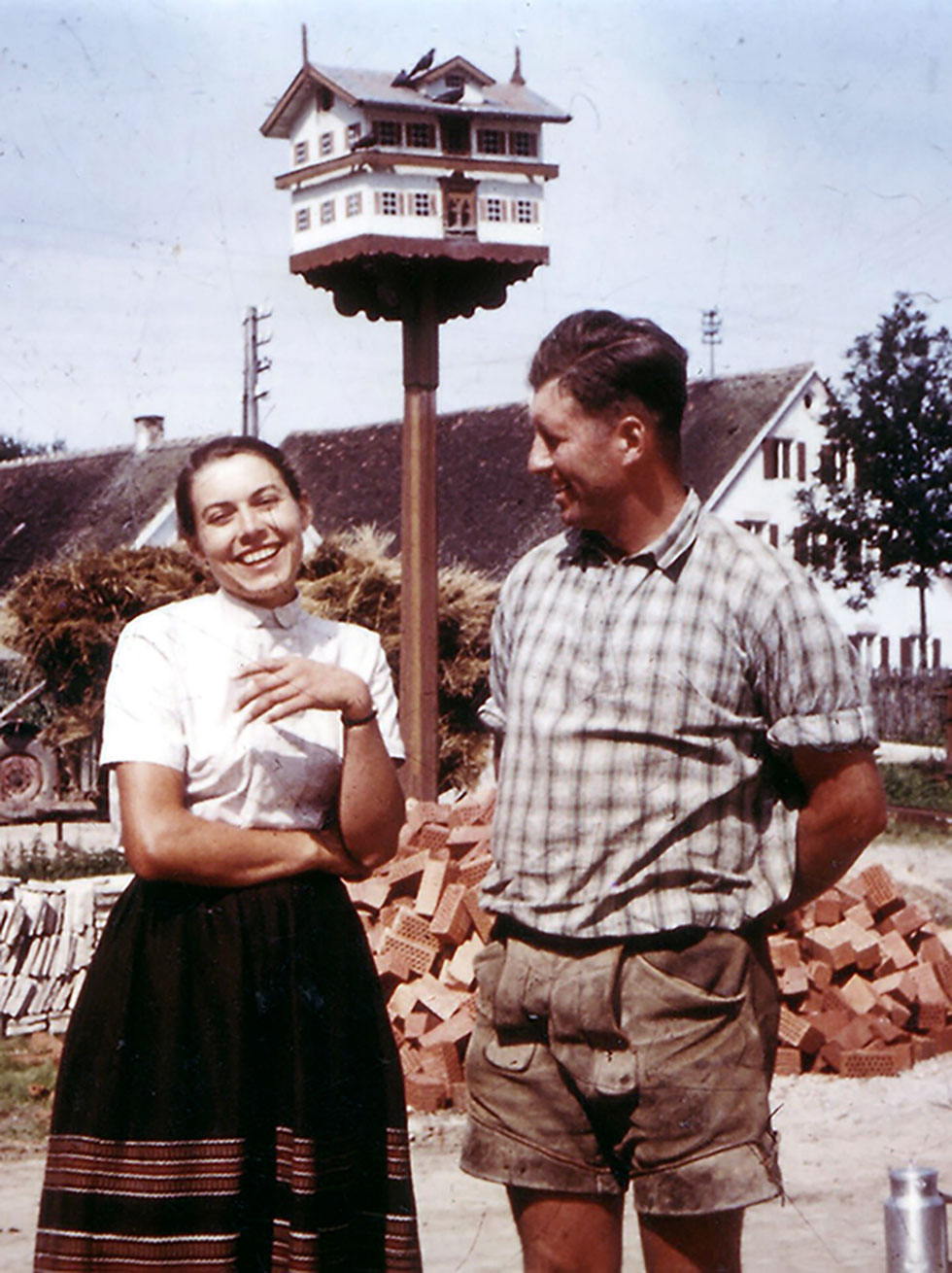 Foto aus den 1950er Jahren: Junges Paar in Arbeitskleidung auf einem Hof mit Taubenschlag und Ziegelhaufen; im Hintergrund Bauernhäuser, wie sie für bayerisch Schwaben typisch waren.