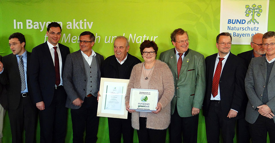 Eine Gruppe Menschen vor einem Bund Naturschutz Hintergrund, teilweise Urkunden in die Kamera Eine Gruppe Menschen vor eienem Bund Naturschutz-Hintergrund. Die beiden Mittleren halten Urkunden der Kamera entgegen.