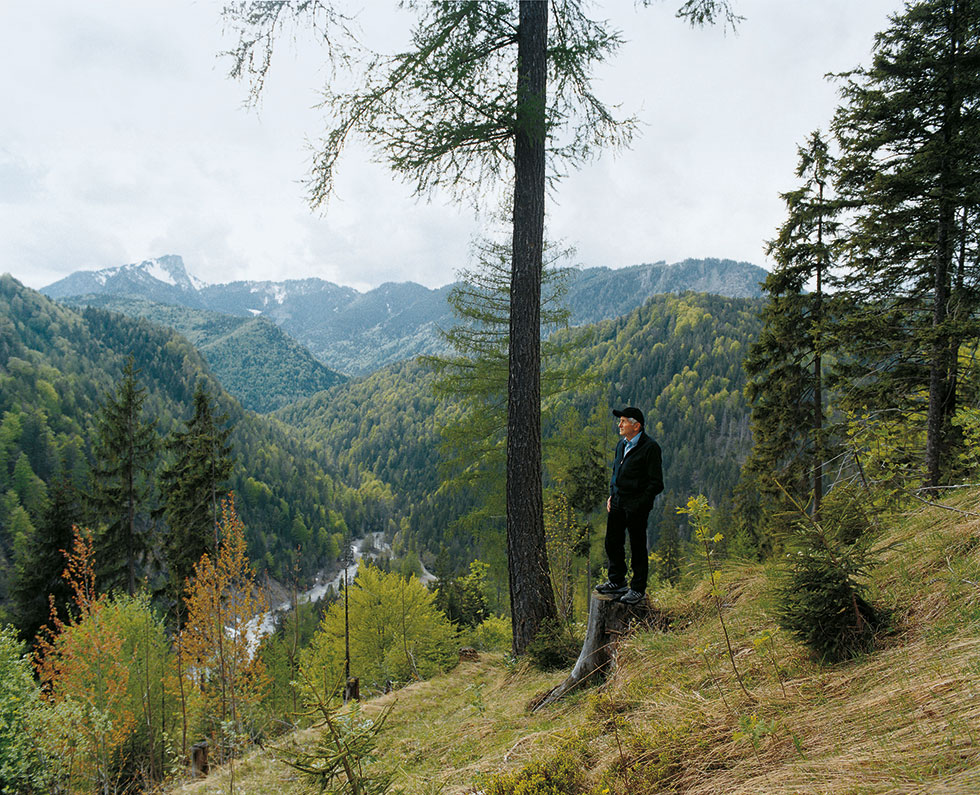 Alois Glück an einem Bergaussichtsplatz auf einem Baumstumpf stehend.