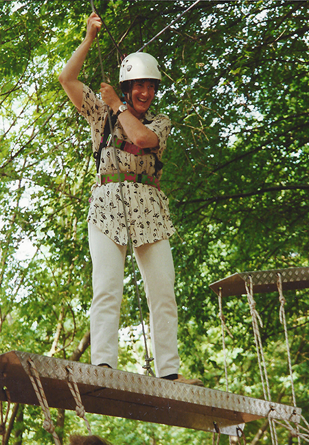 Frau im Tragegeschirr und mit Schutzhelm steht lachend auf einer hölzernen Plattform und hält sich an einem Seil fest.