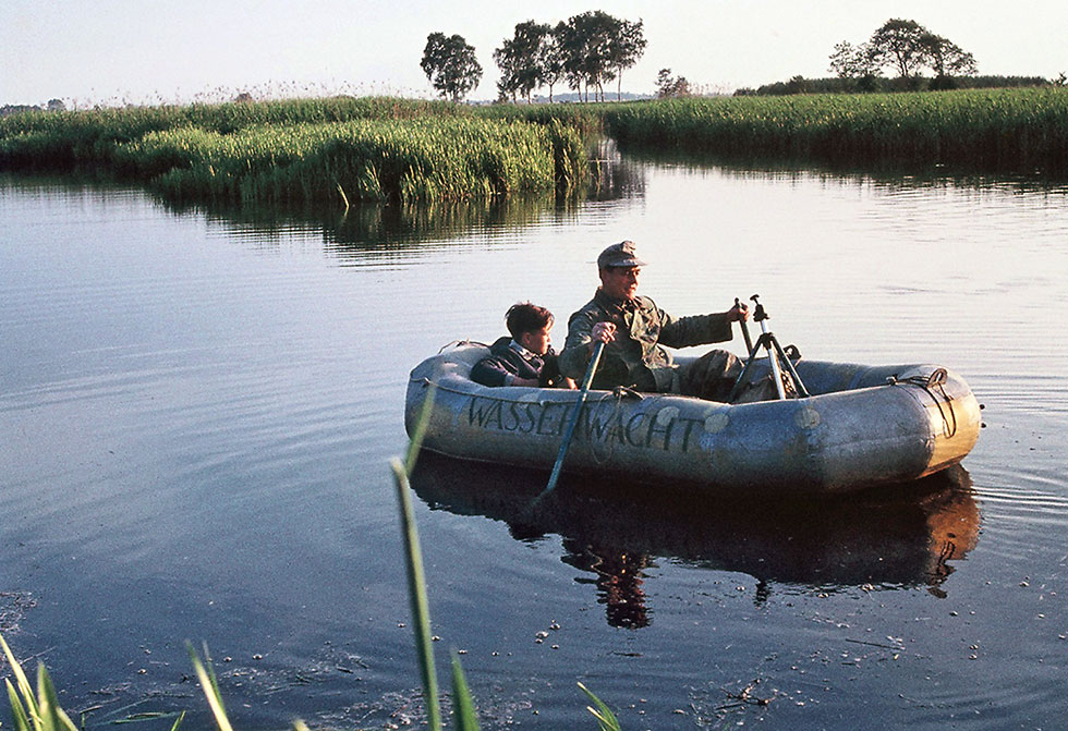 Historisches Foto: Ein Erwachsener und ein Kind in einem Schlauchboot auf einem stillen Gewässer; im Hintergrund flache Schilflandschaft mit einzelnen Bäumen.