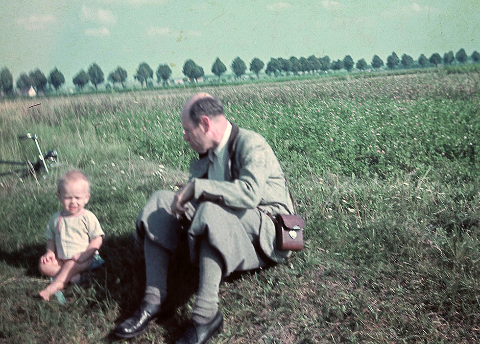 Historisches Foto: Mann und Kleinkind sitzen vor einem ebenen Brachfeld; im Hintergrund verläuft eine Allee.
