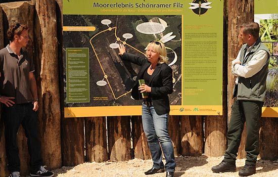 Zwei Männer und eine Frau stehen vor einer Informationstafel. Die Frau erläutert die Tafel.