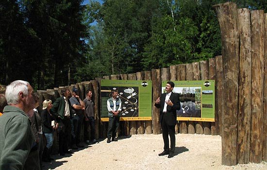 Mehrere Menschen stehen in der Spirale aus stehenden Lärchenstämmen. Ein Mann spricht. Im Hintergrund Wald.