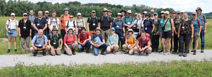 Ein Gruppenfoto mit zirka 40 sommerlich bekleideten Personen im Freien, im Hintergrund ein Auwald