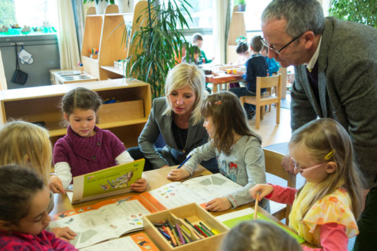 Frau Staatsministerin Scharf kniet am Basteltisch bei den Kindern, die das Entdeckerbuch durchsehen. Der Direktor der ANL, Dieter Pasch, steht dabei und sieht interessiert zu.