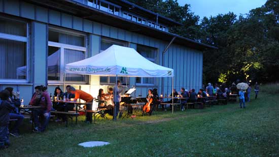 Unter einem Pavillon spielen bei gedämpftem Licht Musiker, während ringsum die Besucher an Tischen sitzen und essen und trinken.