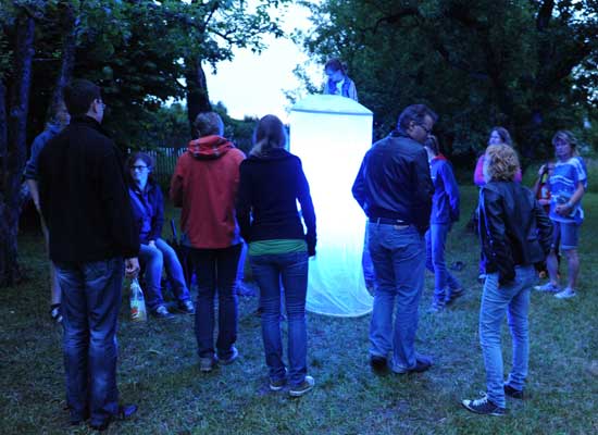 Mehrere Besucher stehen um einen leuchtenden Zylinder, der durch das Licht Insekten anzieht.