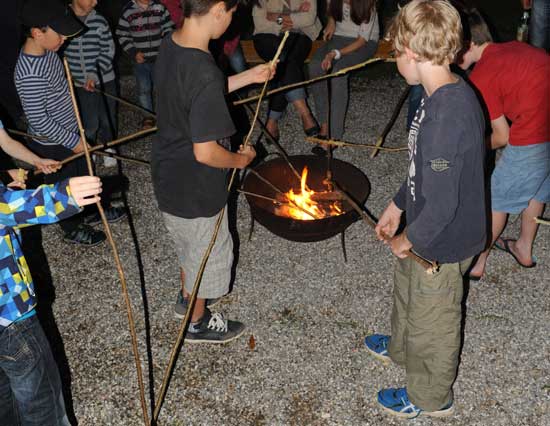 Mehrere Kinder stehen mit Stöcken um eine Feuerschale, um ihre Stockbrote zu backen.